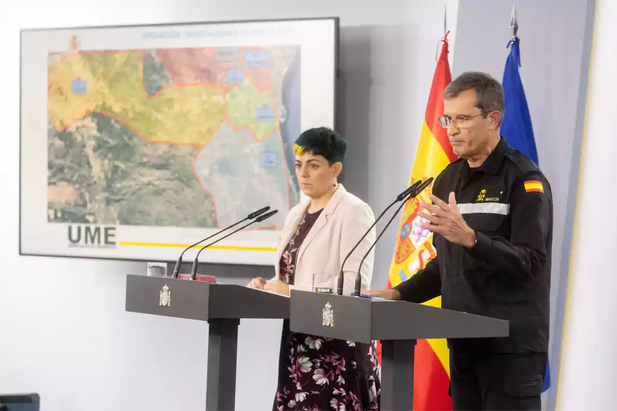 Dos personas en un podio durante una conferencia de prensa con un mapa en el fondo y banderas de España y la Unión Europea.