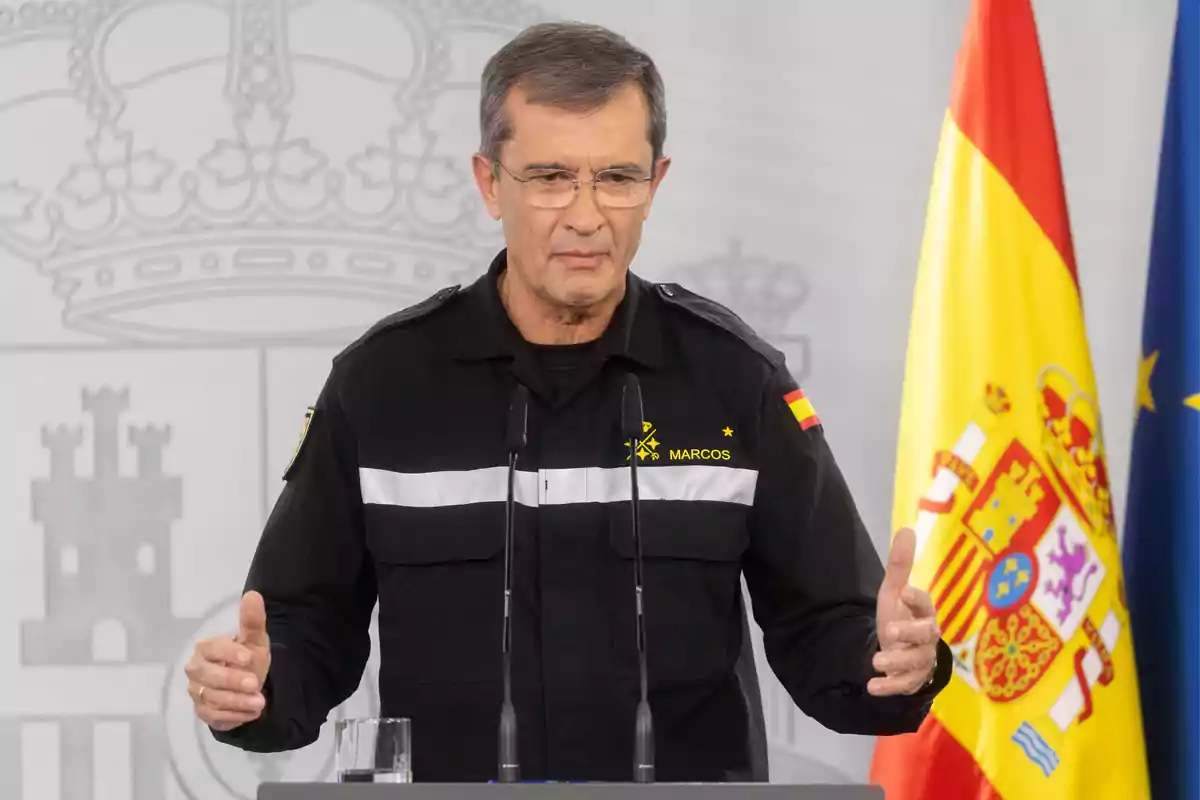 Un hombre con uniforme negro y gafas habla en un podio con banderas de España y la Unión Europea de fondo.