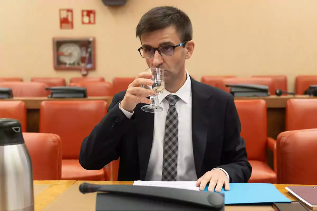 Un hombre con gafas y traje bebe agua de un vaso en una sala de conferencias con sillas rojas.