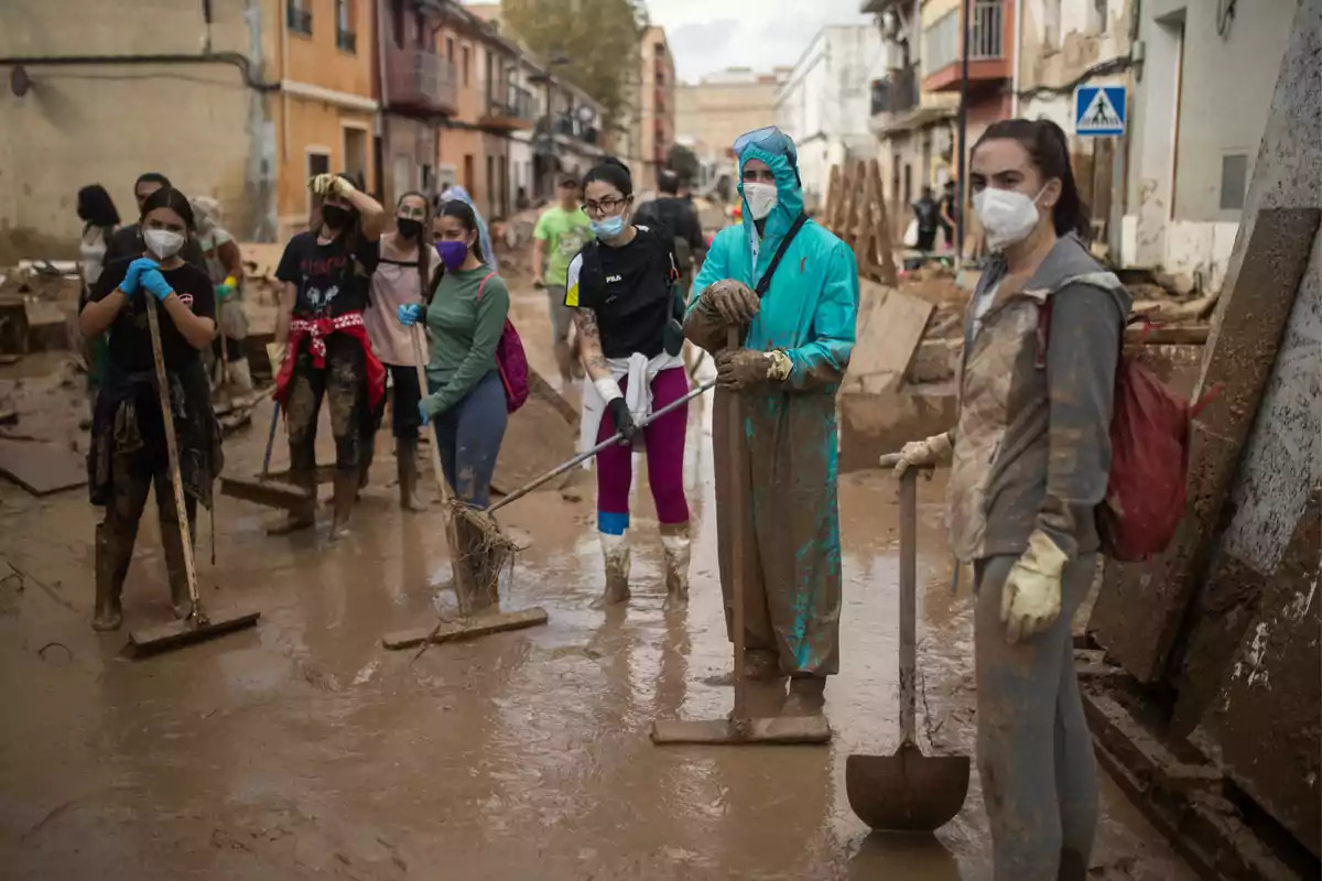 Un grupo de personas con mascarillas y herramientas limpian una calle llena de barro después de una inundación.