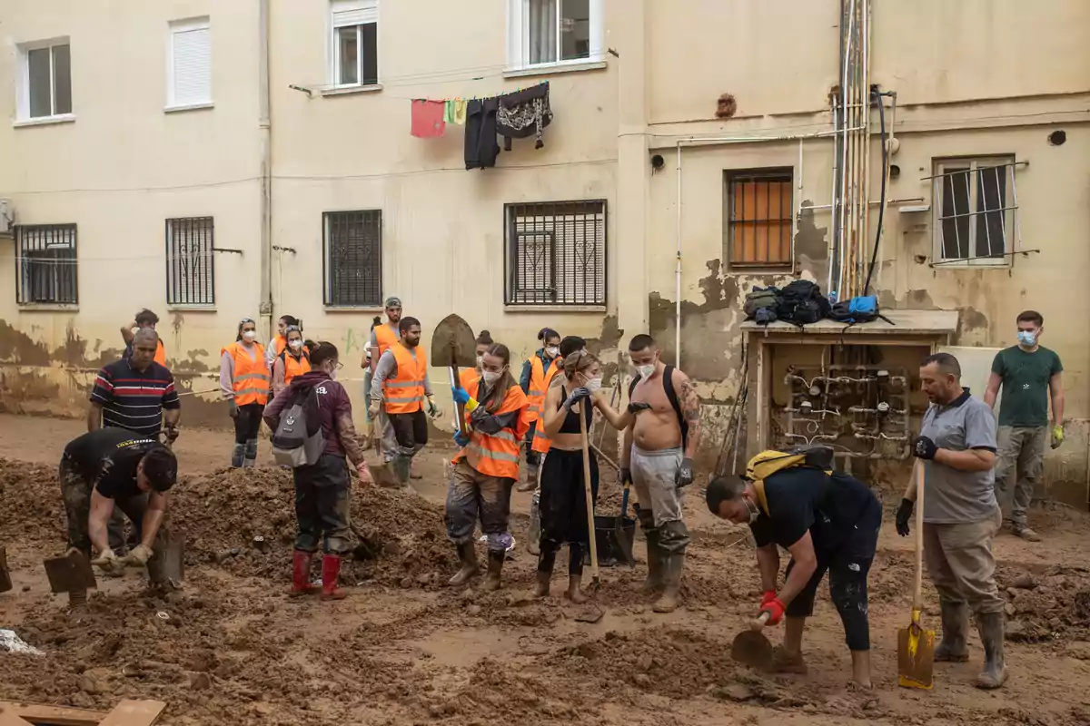 Un grupo de personas con chalecos naranjas y mascarillas trabaja en la limpieza de un área cubierta de barro frente a un edificio.