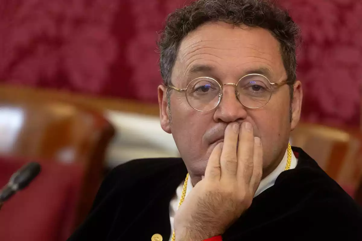 Un hombre con gafas y expresión pensativa se lleva la mano a la boca en un entorno formal.