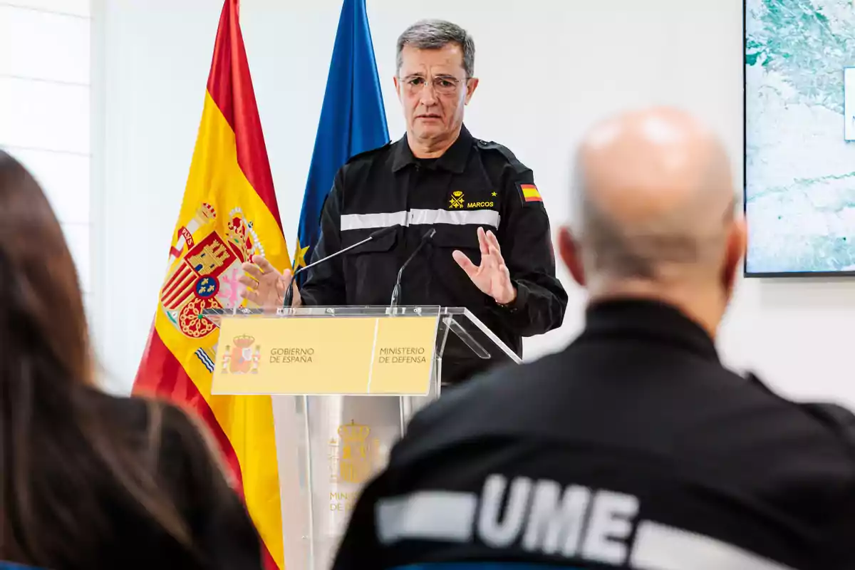 Un hombre en uniforme de la UME habla en un podio con las banderas de España y la Unión Europea detrás.