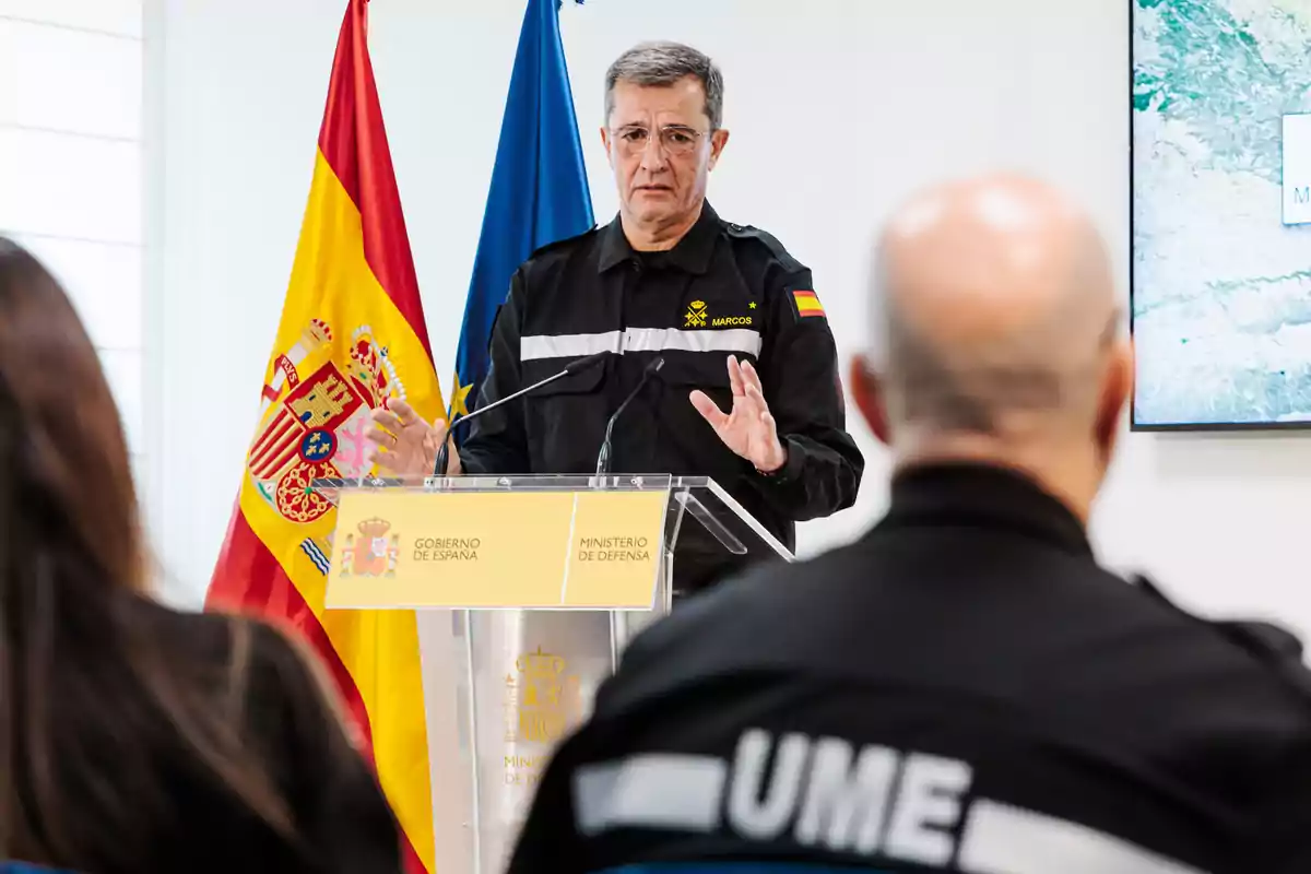 Un hombre en uniforme de la UME habla en un podio con banderas de España y la Unión Europea detrás.