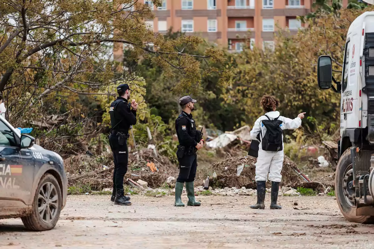 Tres personas, dos policías y una persona con traje blanco, están de pie en un área al aire libre con escombros y vegetación, junto a un vehículo policial y otro vehículo.