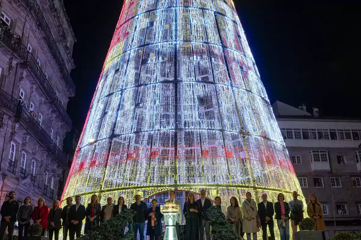 Un grupo de personas posa frente a un gran árbol de Navidad iluminado con luces de colores en una plaza urbana por la noche.