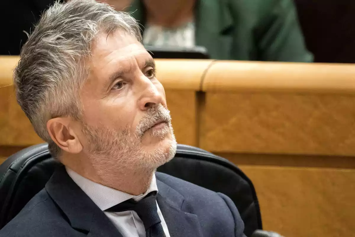 Un hombre de cabello canoso y barba está sentado en un entorno formal con expresión pensativa.