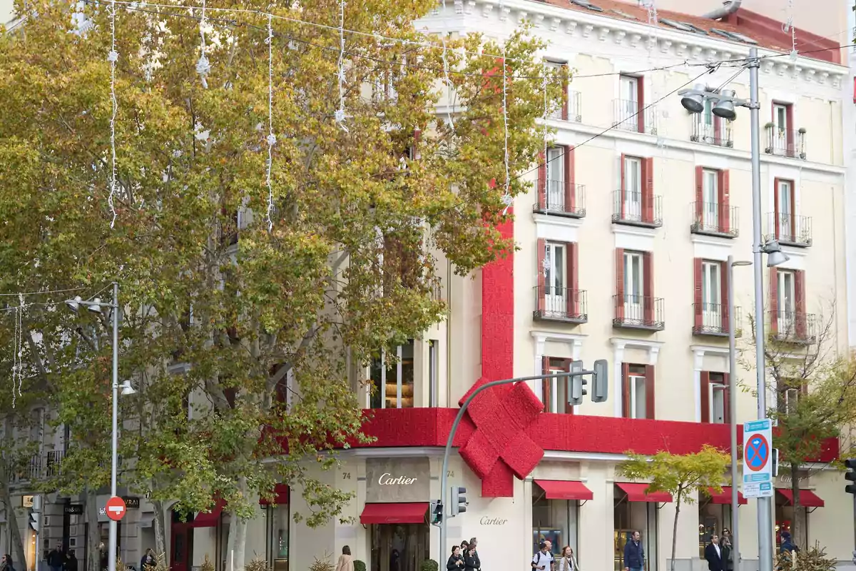 Fachada de una tienda Cartier decorada con un gran lazo rojo, rodeada de árboles y personas caminando por la acera.