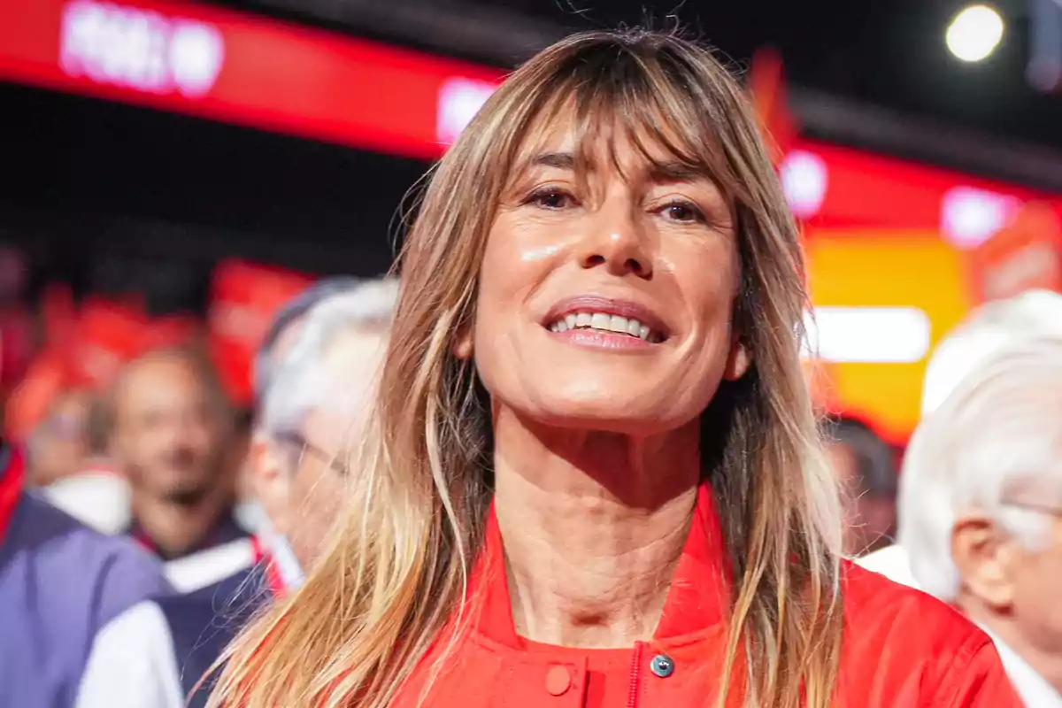 Una mujer sonriente con chaqueta roja en un evento con luces y personas de fondo.