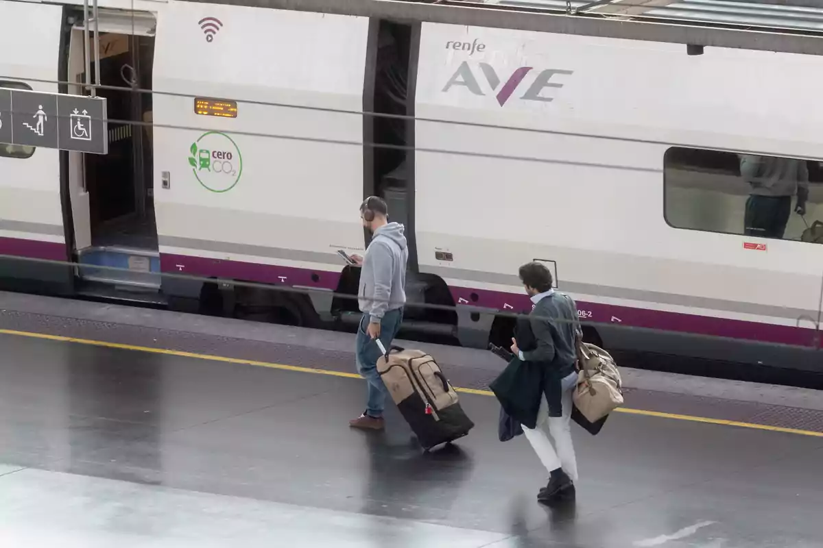 Dos personas caminando con maletas junto a un tren de alta velocidad en una estación.