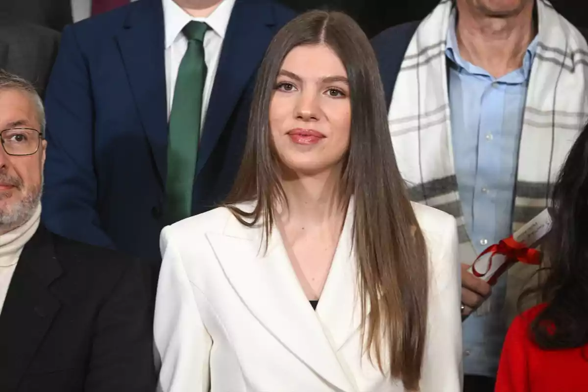 Una mujer con cabello largo y lacio lleva un traje blanco y está rodeada de otras personas en un evento formal. Una mujer con cabello largo y lacio lleva un traje blanco y está rodeada de otras personas en un evento formal.