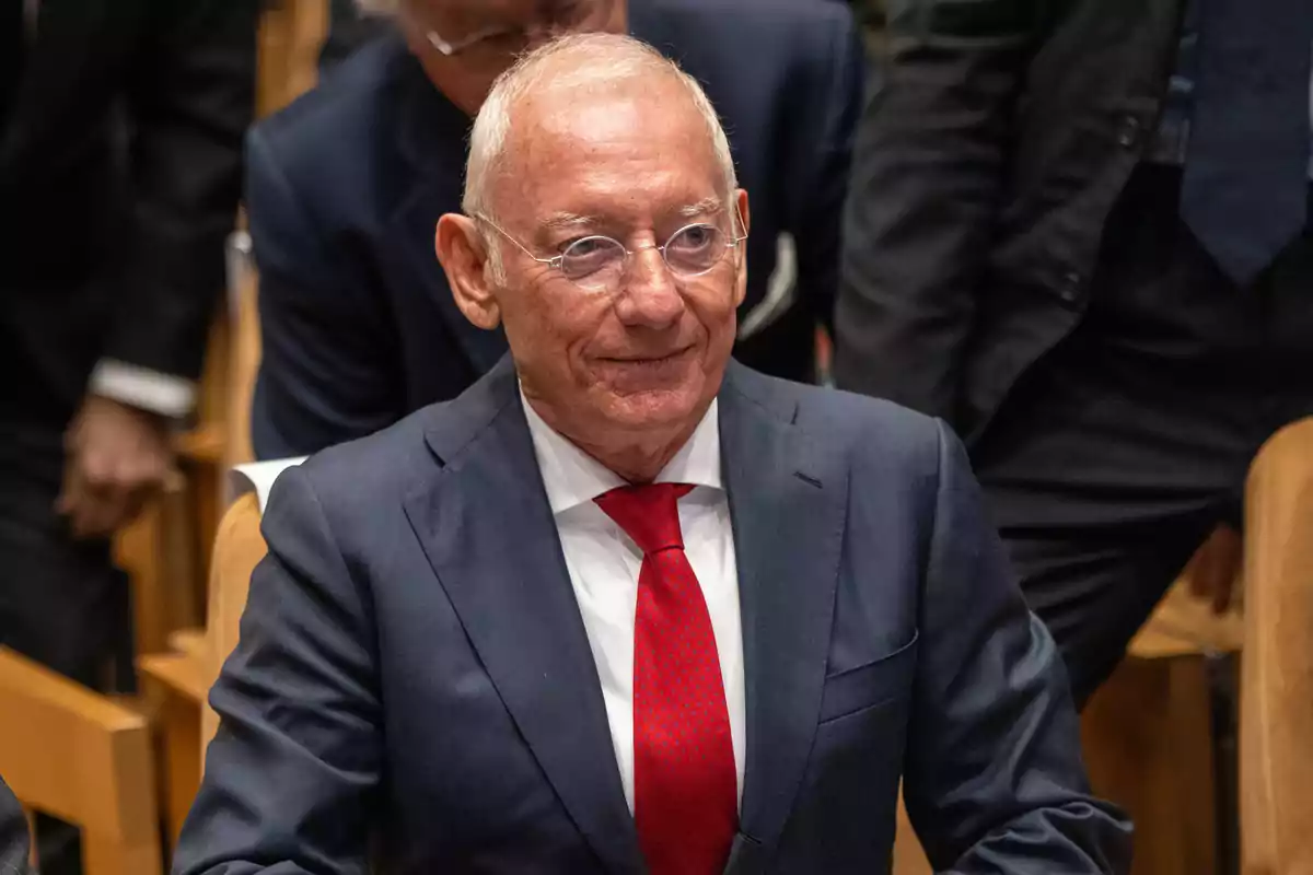 Un hombre mayor con gafas y traje oscuro con corbata roja está sentado en un auditorio.