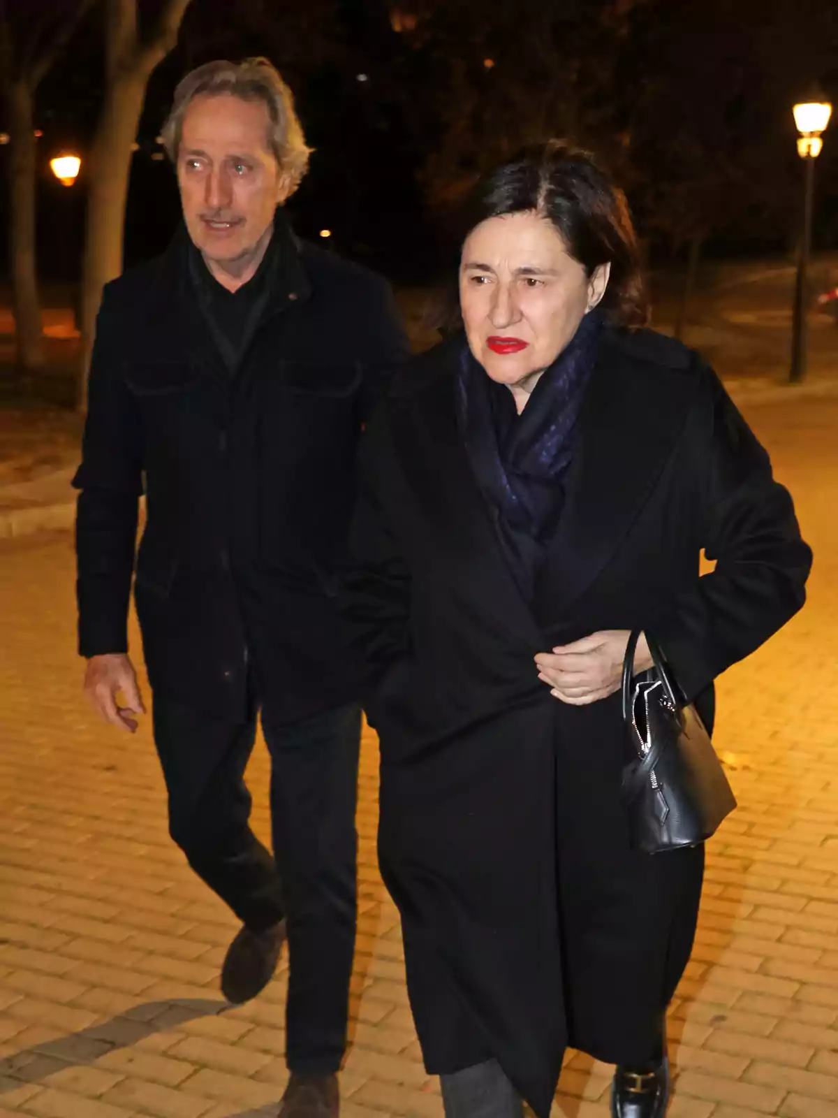 A woman and a man walk together down a street lit by streetlights at night, both wearing dark coats and the woman holding a black purse. A woman and a man walk together down a street lit by streetlights at night, both wearing dark coats and the woman holding a black purse.