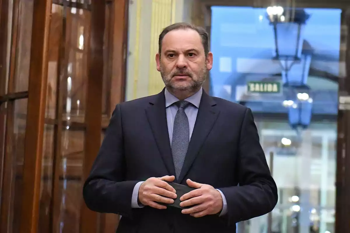 Hombre de mediana edad con barba y traje oscuro de pie en un pasillo elegante con un letrero de salida al fondo