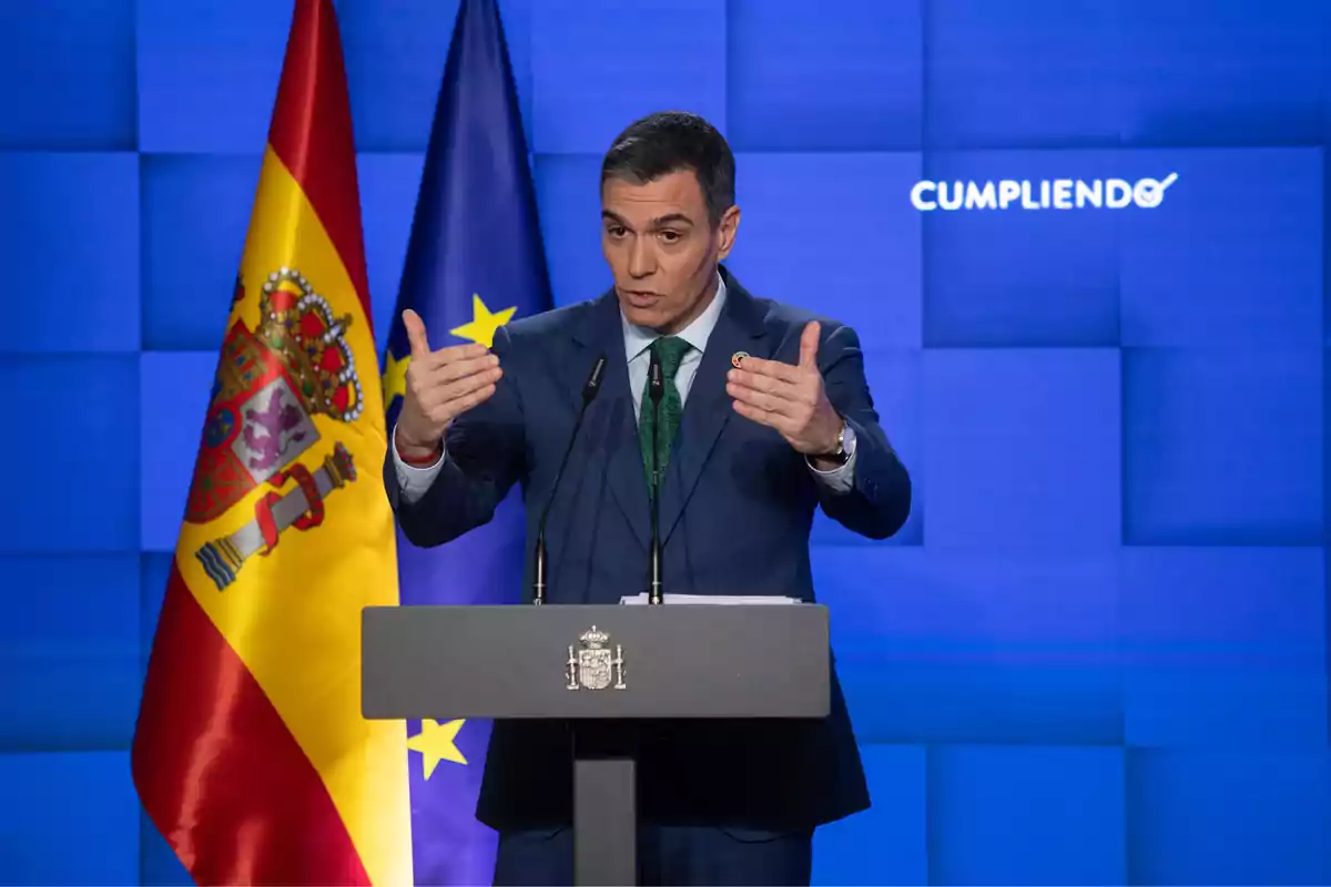 Un hombre en traje azul habla en un podio con banderas de España y la Unión Europea detrás.