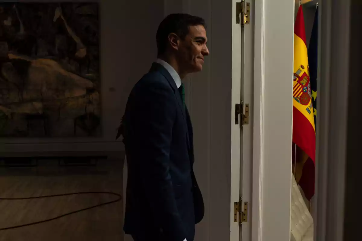 Un hombre de traje oscuro está de perfil junto a una puerta abierta, con una bandera de España visible al fondo.