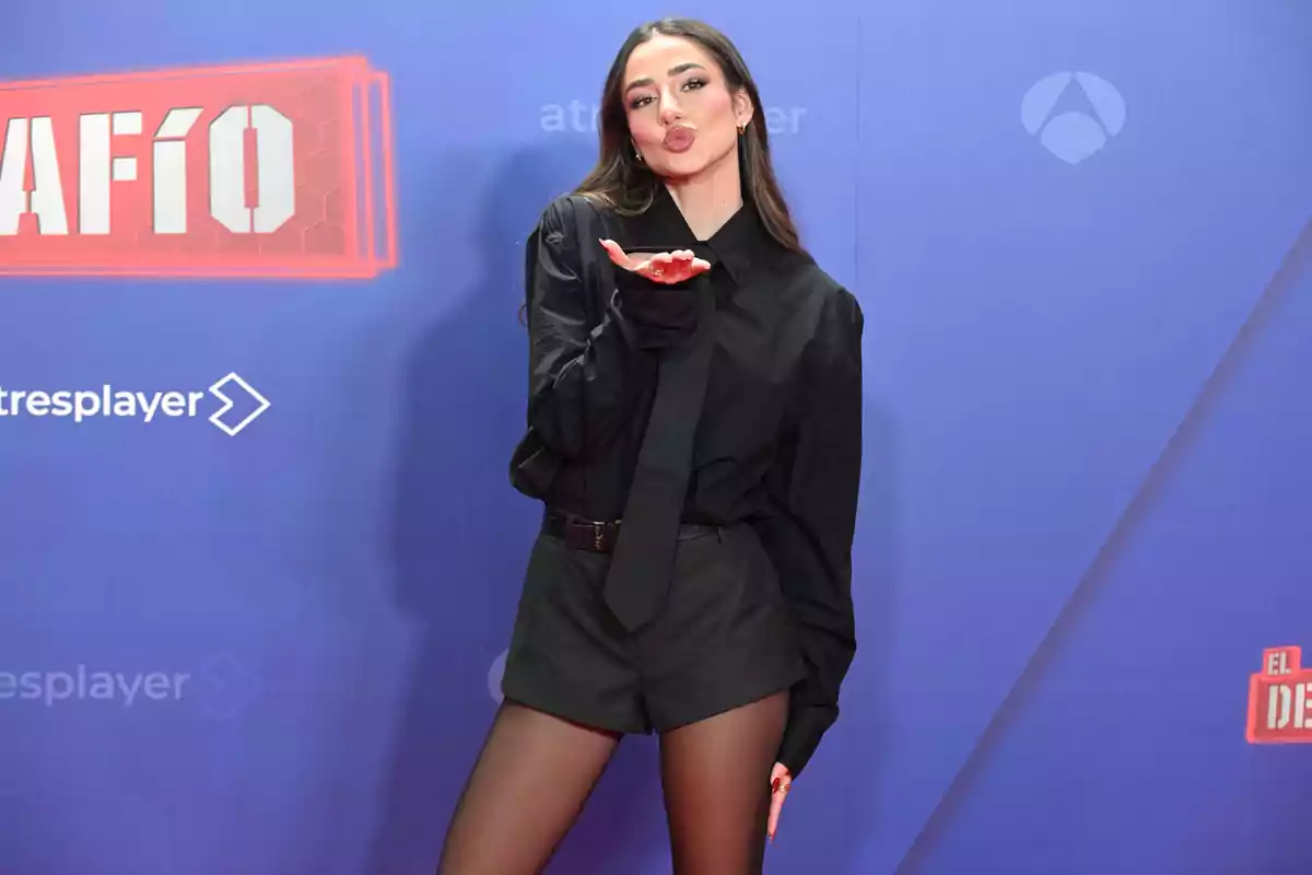 Mujer posando en una alfombra roja con fondo azul, vestida con camisa negra, corbata, shorts y medias, lanzando un beso hacia la cámara Mujer posando en una alfombra roja con fondo azul, vestida con camisa negra, corbata, shorts y medias, lanzando un beso hacia la cámara