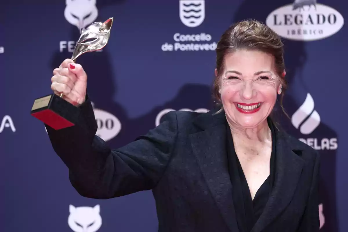 Una persona sonriente sostiene un trofeo en un evento con fondo azul.