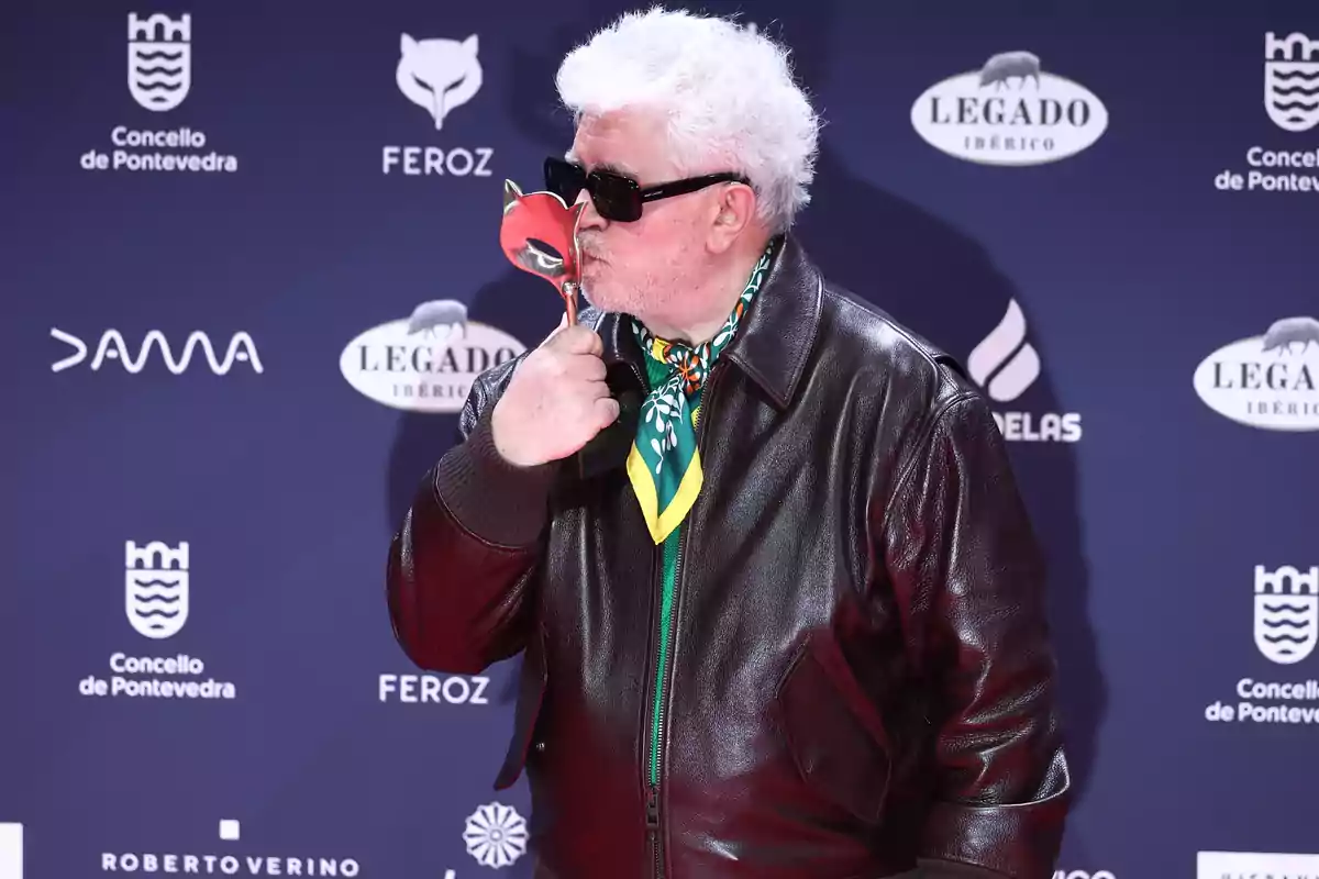 Un hombre con gafas de sol y chaqueta de cuero besa un trofeo en un evento con fondo de logotipos.