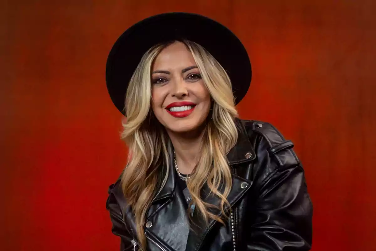 Mujer con sombrero negro y chaqueta de cuero sonriendo frente a un fondo rojo.