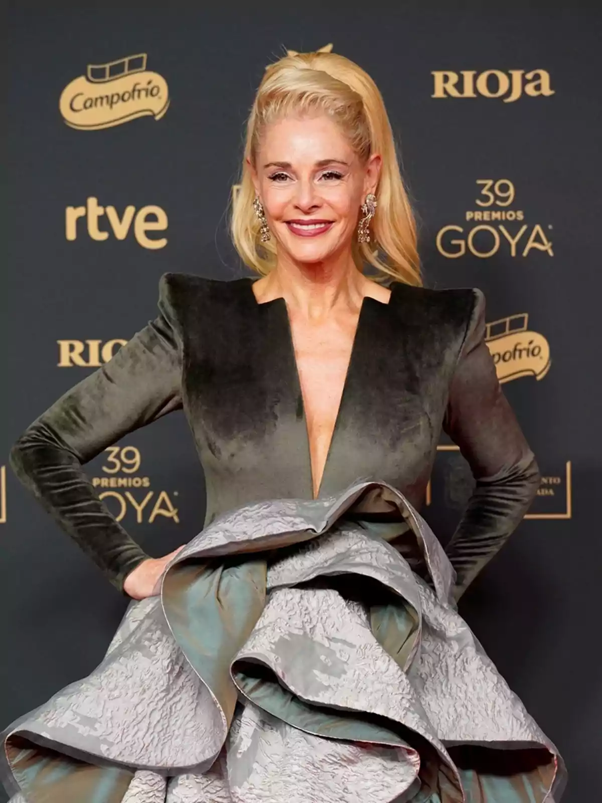 Mujer rubia con vestido elegante de terciopelo verde y falda voluminosa gris posando sonriente en la alfombra roja de los Premios Goya Mujer rubia con vestido elegante de terciopelo verde y falda voluminosa gris posando sonriente en la alfombra roja de los Premios Goya