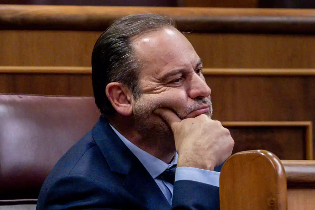 Hombre de mediana edad con traje azul sentado en un escaño de madera apoyando la barbilla en su mano y mirando pensativo hacia un lado