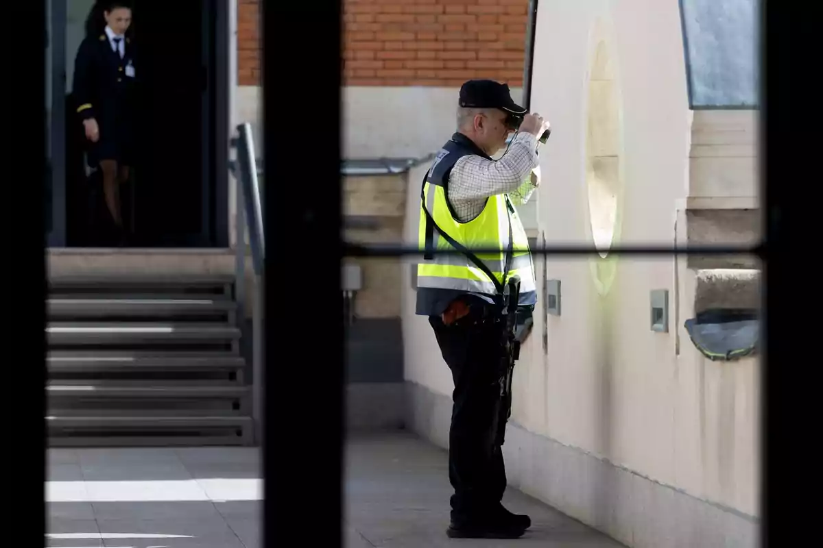 Un guardia de seguridad con chaleco amarillo observa a través de un visor mientras una persona uniformada baja unas escaleras en el fondo. Un guardia de seguridad con chaleco amarillo observa a través de un visor mientras una persona uniformada baja unas escaleras en el fondo.