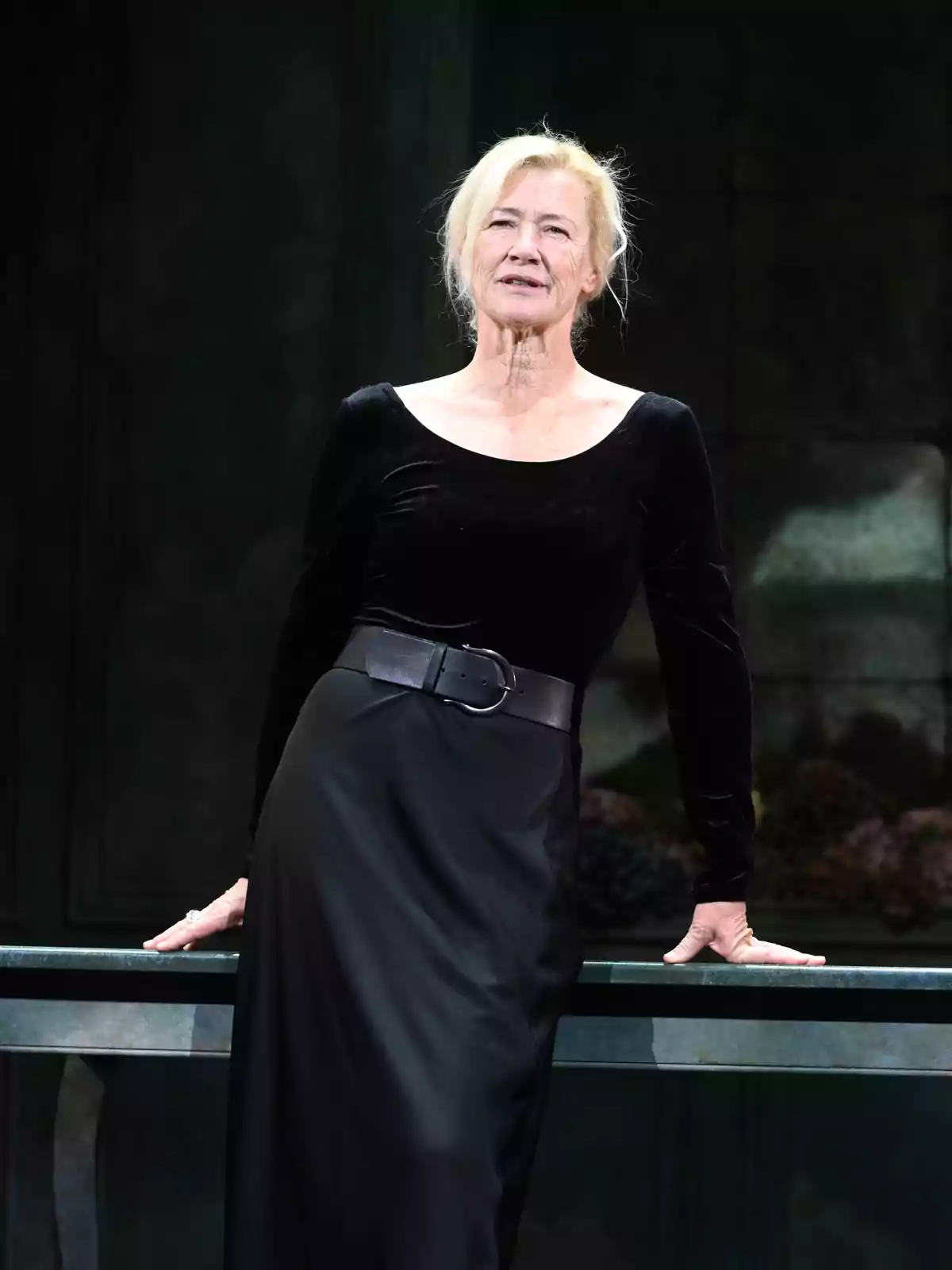 Mujer de cabello rubio recogido vestida con un vestido negro elegante apoyada sobre una mesa en un escenario teatral Mujer de cabello rubio recogido vestida con un vestido negro elegante apoyada sobre una mesa en un escenario teatral