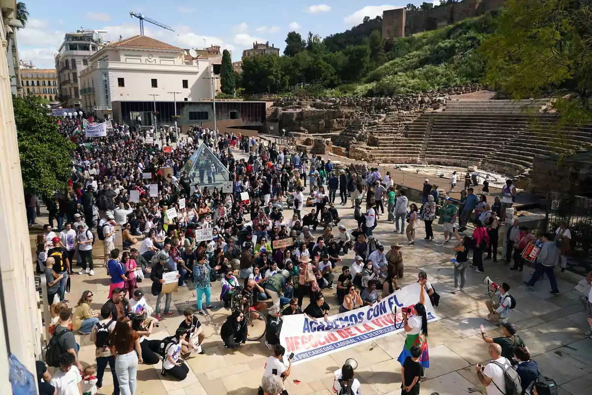 Una multitud de personas se reúne en una plaza al aire libre, algunas sosteniendo pancartas, con un antiguo teatro romano al fondo y edificios modernos alrededor. Una multitud de personas se reúne en una plaza al aire libre, algunas sosteniendo pancartas, con un antiguo teatro romano al fondo y edificios modernos alrededor.