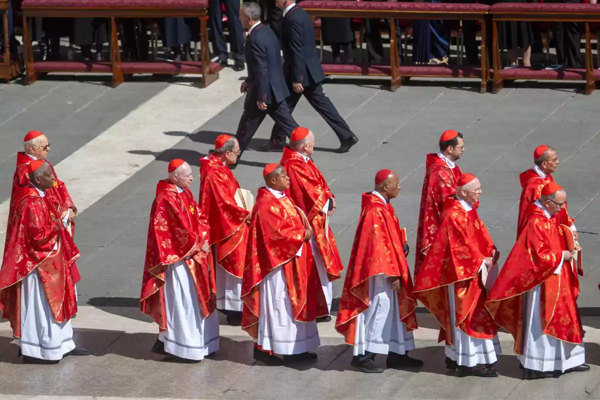 Un grupo de cardenales vestidos con sotanas rojas camina en fila durante una ceremonia al aire libre. Un grupo de cardenales vestidos con sotanas rojas camina en fila durante una ceremonia al aire libre.