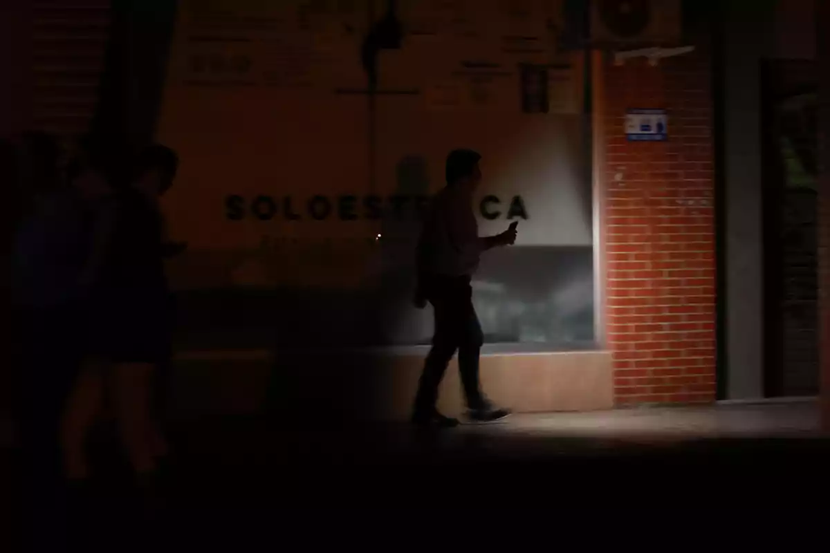 Una persona camina por una calle oscura sosteniendo un teléfono móvil, con un fondo de ladrillos y un letrero parcialmente visible. Una persona camina por una calle oscura sosteniendo un teléfono móvil, con un fondo de ladrillos y un letrero parcialmente visible.