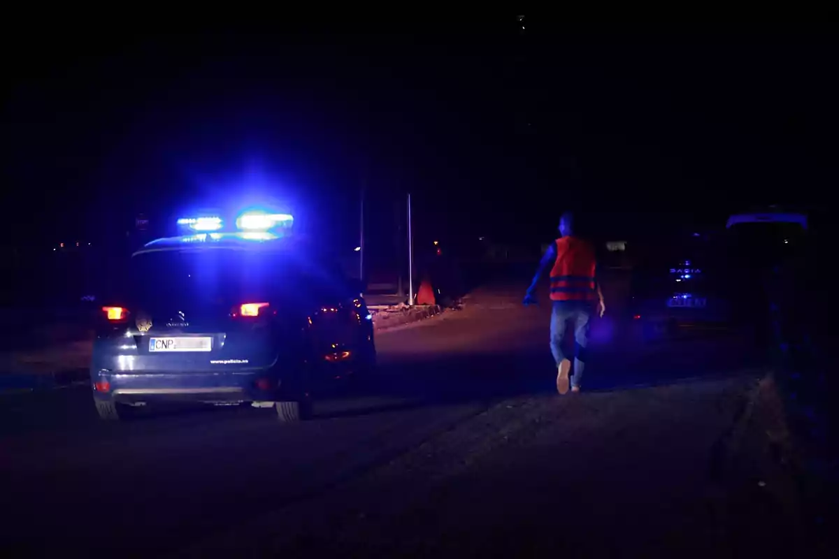 Una escena nocturna muestra un coche de policía con luces azules encendidas y una persona con chaleco reflectante caminando por la carretera. Una escena nocturna muestra un coche de policía con luces azules encendidas y una persona con chaleco reflectante caminando por la carretera.