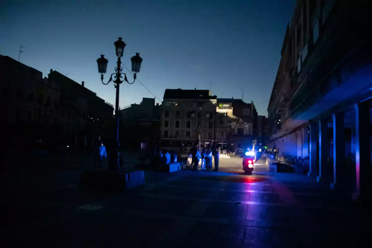 Una plaza oscura al anochecer con un farol y luces de un vehículo iluminando a un grupo de personas. Una plaza oscura al anochecer con un farol y luces de un vehículo iluminando a un grupo de personas.