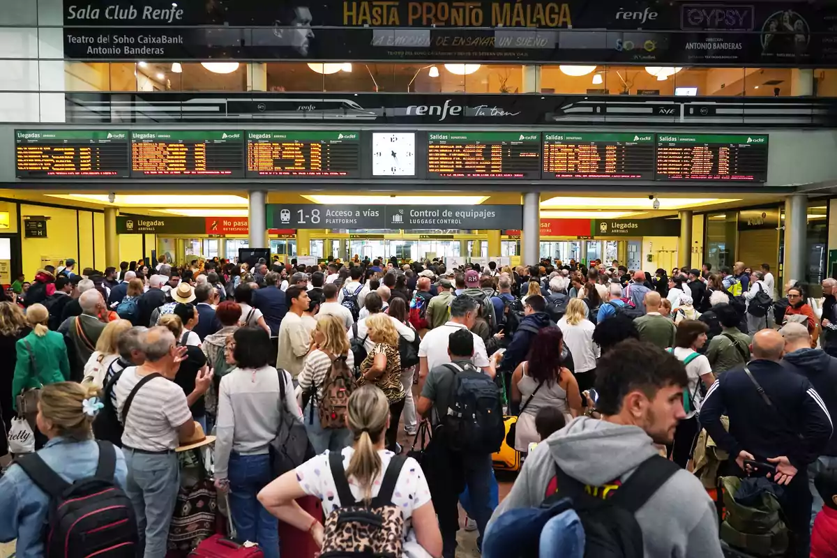 Un grupo numeroso de personas se encuentra reunido en una estación de tren, observando los paneles de información de llegadas y salidas.