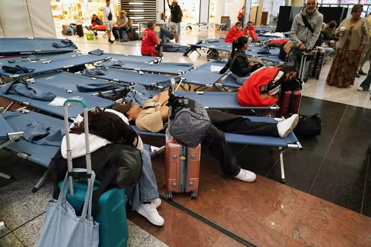 Personas descansando en camillas en un área de espera de un aeropuerto con equipaje a su alrededor. Personas descansando en camillas en un área de espera de un aeropuerto con equipaje a su alrededor.