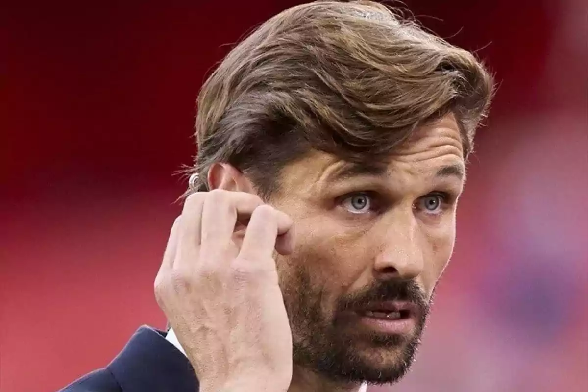Hombre de cabello castaño y barba con expresión seria ajustando un auricular en su oído sobre un fondo rojo difuminado Hombre de cabello castaño y barba con expresión seria ajustando un auricular en su oído sobre un fondo rojo difuminado