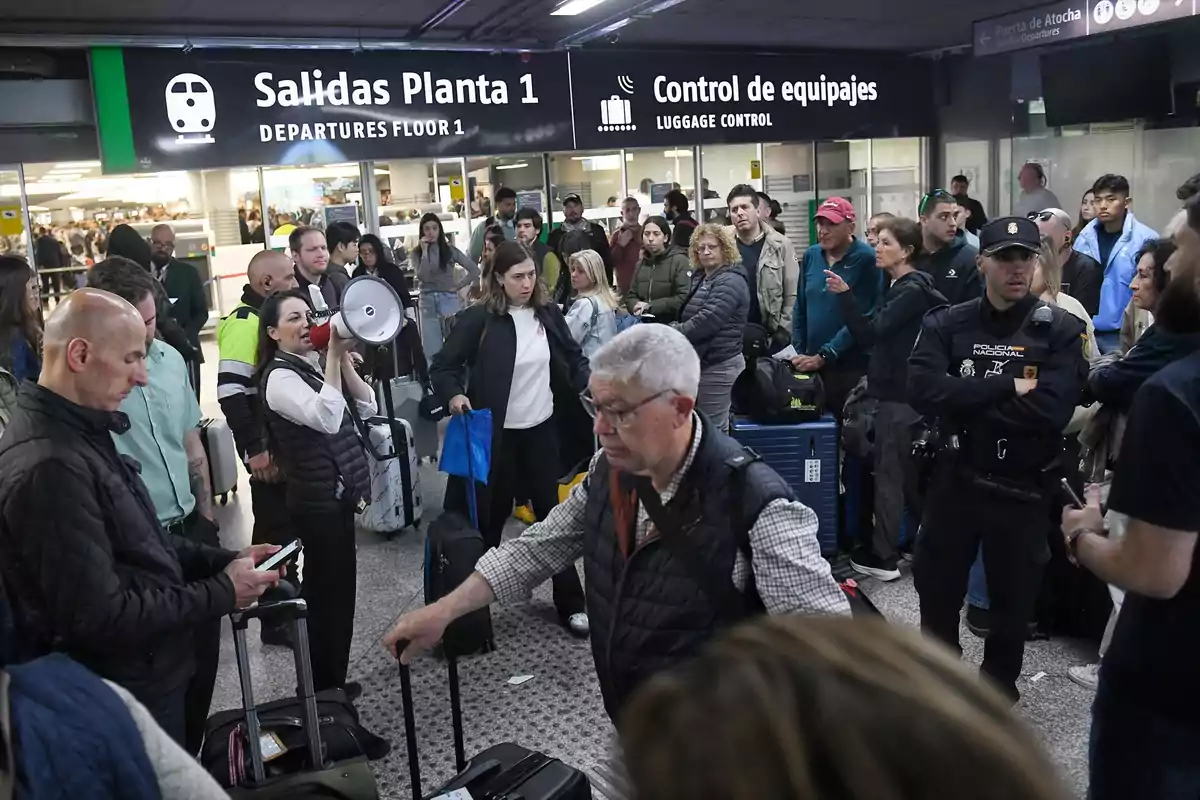 Un grupo de personas se encuentra reunido en un área de salidas de un aeropuerto, algunas con equipaje, mientras una mujer habla por un megáfono y un policía observa la escena. Un grupo de personas se encuentra reunido en un área de salidas de un aeropuerto, algunas con equipaje, mientras una mujer habla por un megáfono y un policía observa la escena.