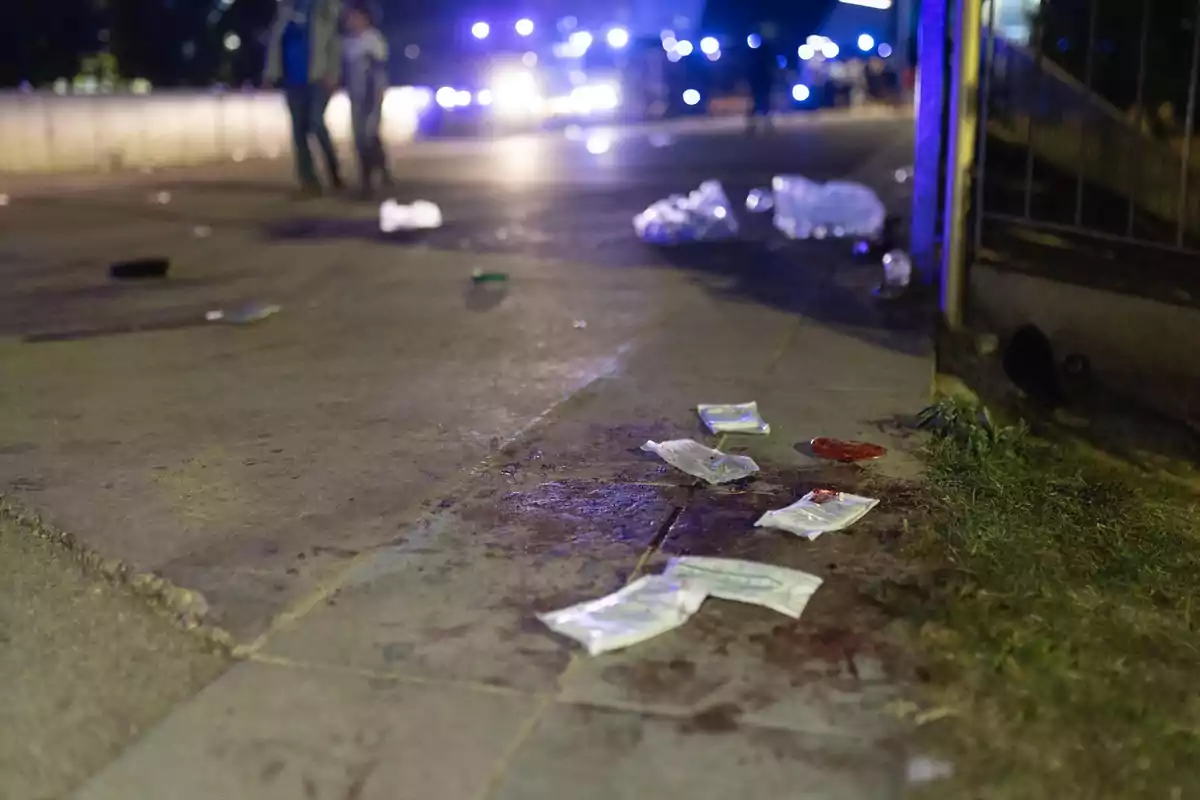 Una escena nocturna muestra una acera con manchas oscuras y bolsas de plástico esparcidas, mientras luces azules brillan en el fondo y dos personas caminan a lo lejos. Una escena nocturna muestra una acera con manchas oscuras y bolsas de plástico esparcidas, mientras luces azules brillan en el fondo y dos personas caminan a lo lejos.