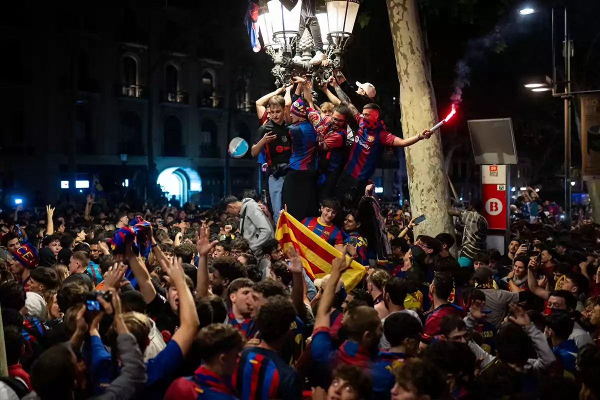 Un grupo de personas celebra en la calle, algunas subidas a un poste de luz, con banderas y bengalas, rodeadas de una multitud. Un grupo de personas celebra en la calle, algunas subidas a un poste de luz, con banderas y bengalas, rodeadas de una multitud.