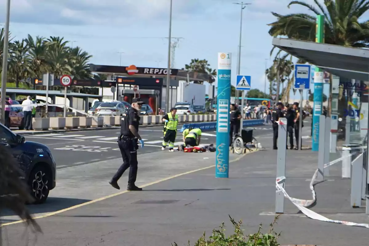 Un grupo de policías y personal de emergencia atienden una situación en una calle cerca de una estación de servicio, con una persona en el suelo y una silla de ruedas cercana. Un grupo de policías y personal de emergencia atienden una situación en una calle cerca de una estación de servicio, con una persona en el suelo y una silla de ruedas cercana.