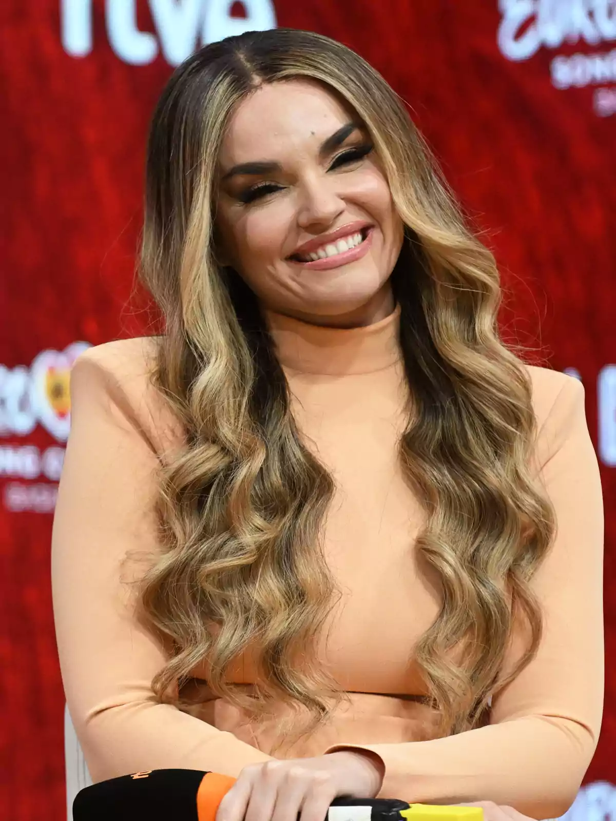 Mujer de cabello largo y ondulado con vestido color durazno sonriendo frente a un fondo rojo Mujer de cabello largo y ondulado con vestido color durazno sonriendo frente a un fondo rojo