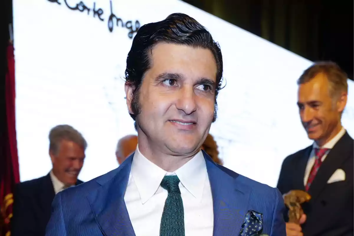 Hombre de cabello oscuro y peinado hacia atrás vistiendo un traje azul con corbata verde en un evento formal con personas al fondo Hombre de cabello oscuro y peinado hacia atrás vistiendo un traje azul con corbata verde en un evento formal con personas al fondo