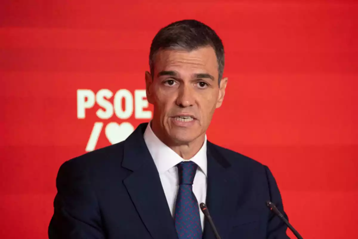 Hombre de cabello corto y canoso con traje azul y corbata hablando frente a un fondo rojo con el logo de PSOE Hombre de cabello corto y canoso con traje azul y corbata hablando frente a un fondo rojo con el logo de PSOE