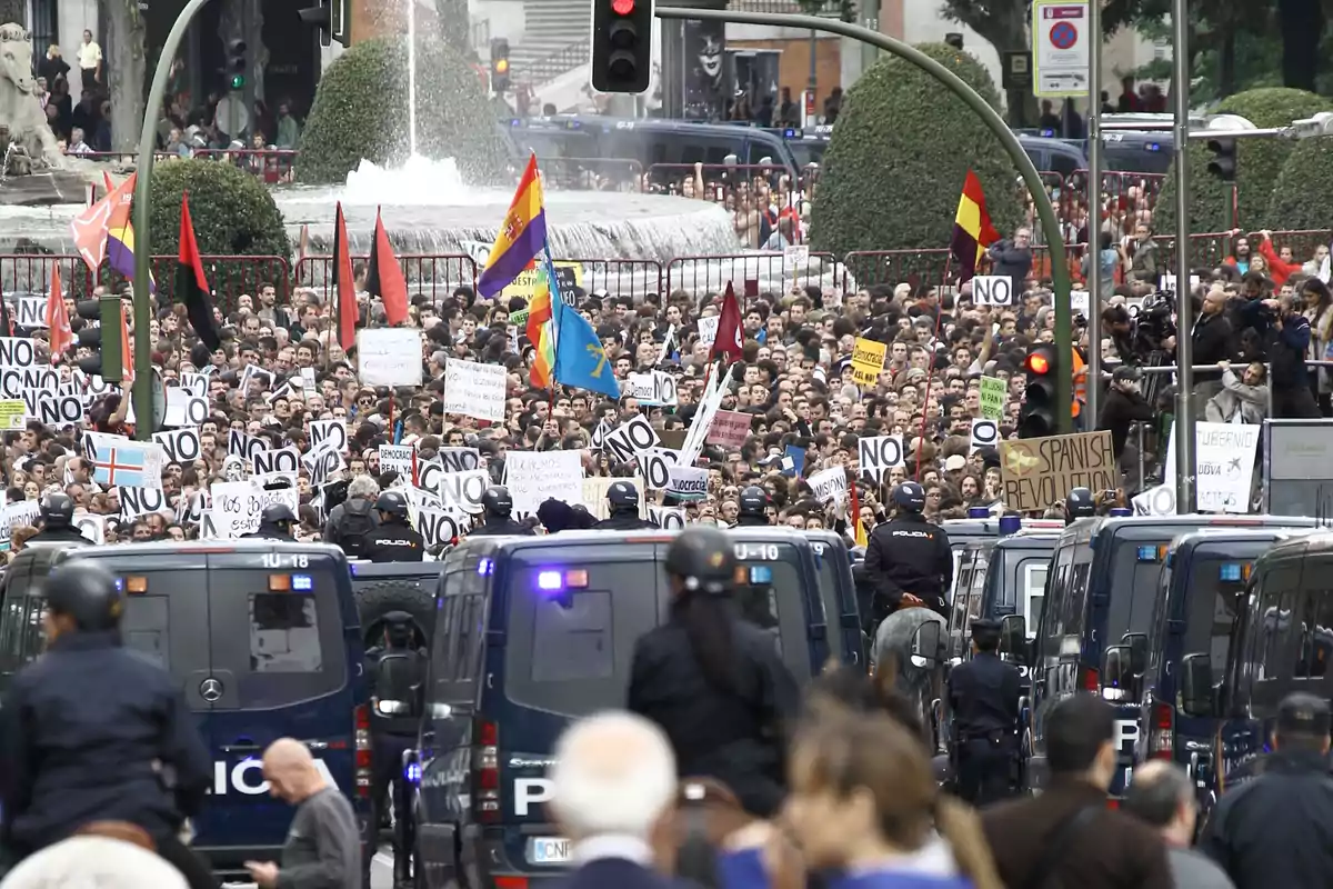 Una multitud de personas se manifiesta en una calle, sosteniendo pancartas y banderas, mientras la policía observa desde vehículos. Una multitud de personas se manifiesta en una calle, sosteniendo pancartas y banderas, mientras la policía observa desde vehículos.