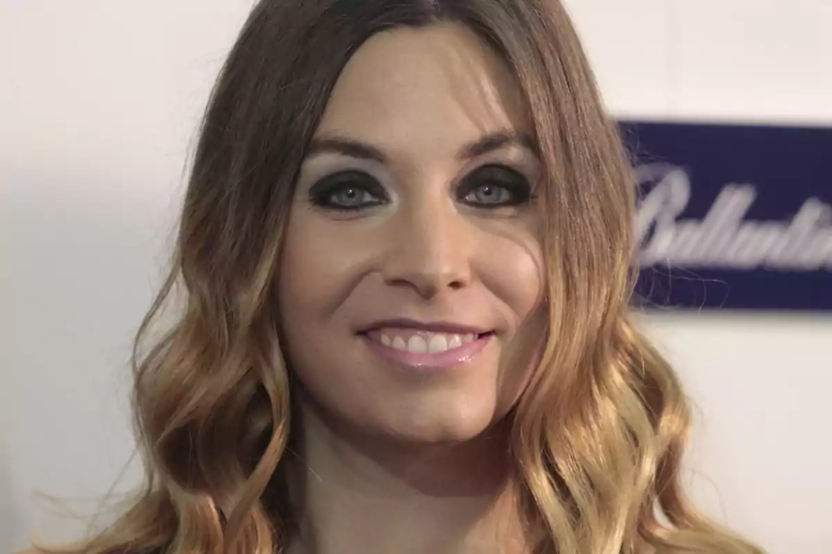 Una mujer con cabello largo y ondulado, maquillaje de ojos oscuro y una sonrisa, con un fondo borroso que incluye un letrero azul.