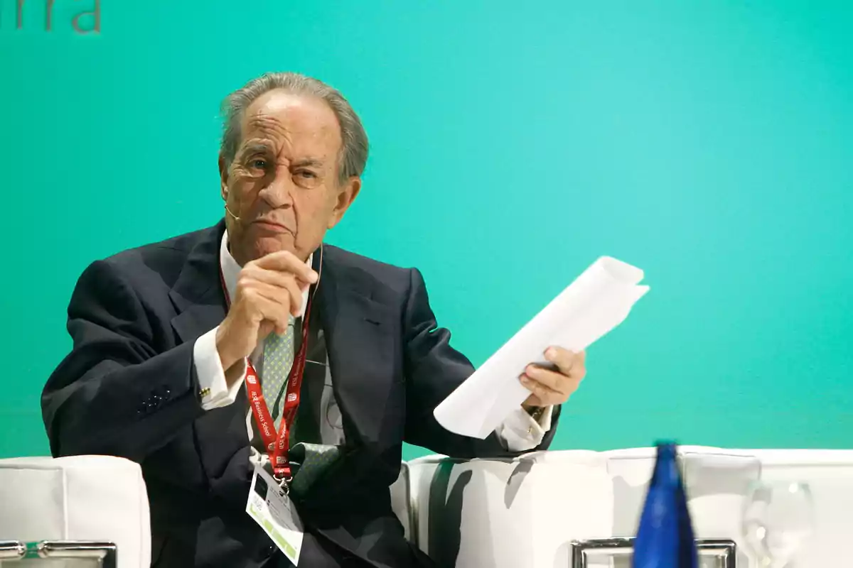 Hombre mayor en traje sosteniendo papeles durante una conferencia con fondo verde.