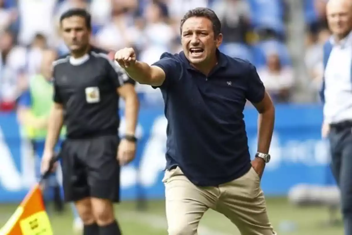 Un entrenador de fútbol gesticula enérgicamente durante un partido mientras un árbitro asiste en el fondo.