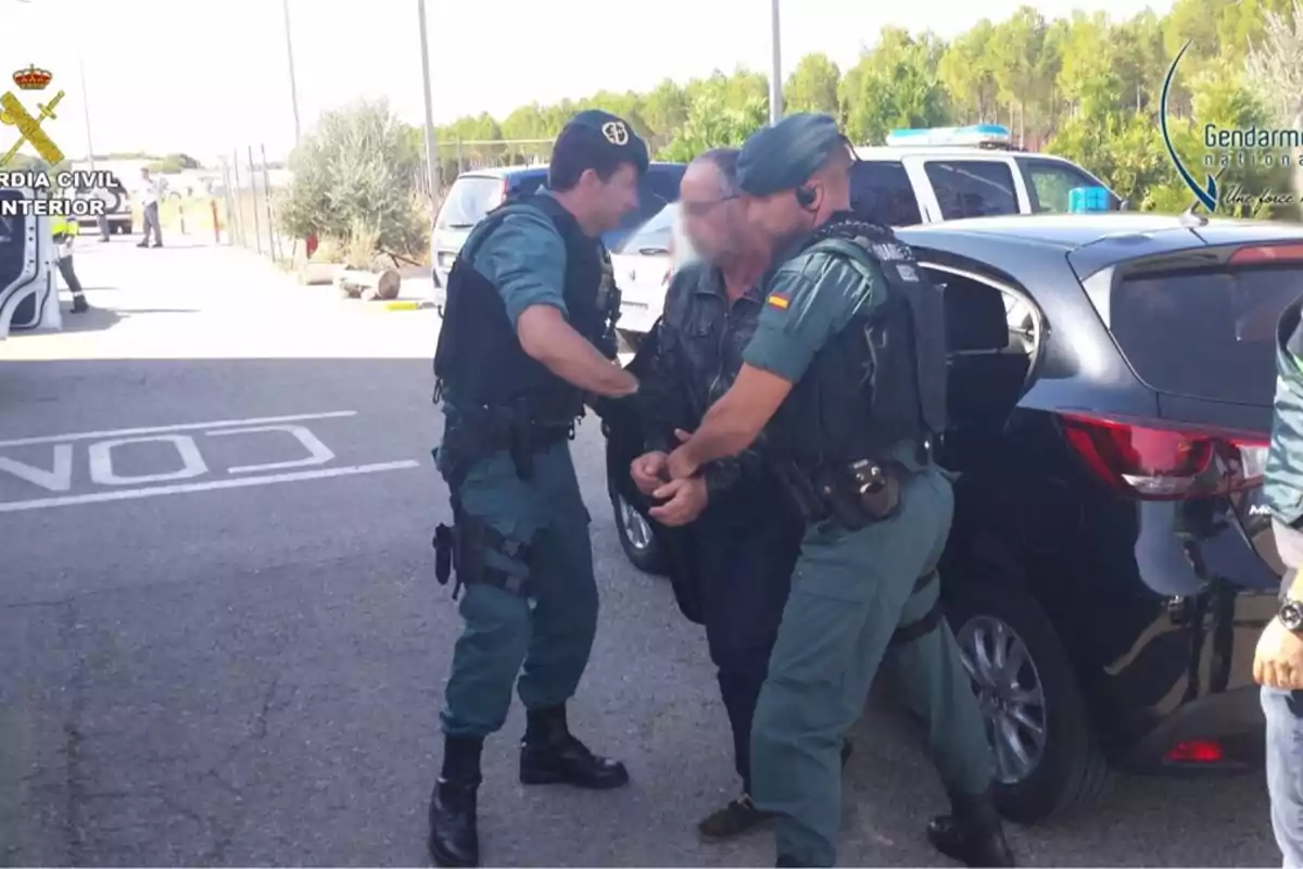 Dos agentes de la Guardia Civil escoltan a un hombre esposado hacia un vehículo en un área al aire libre.