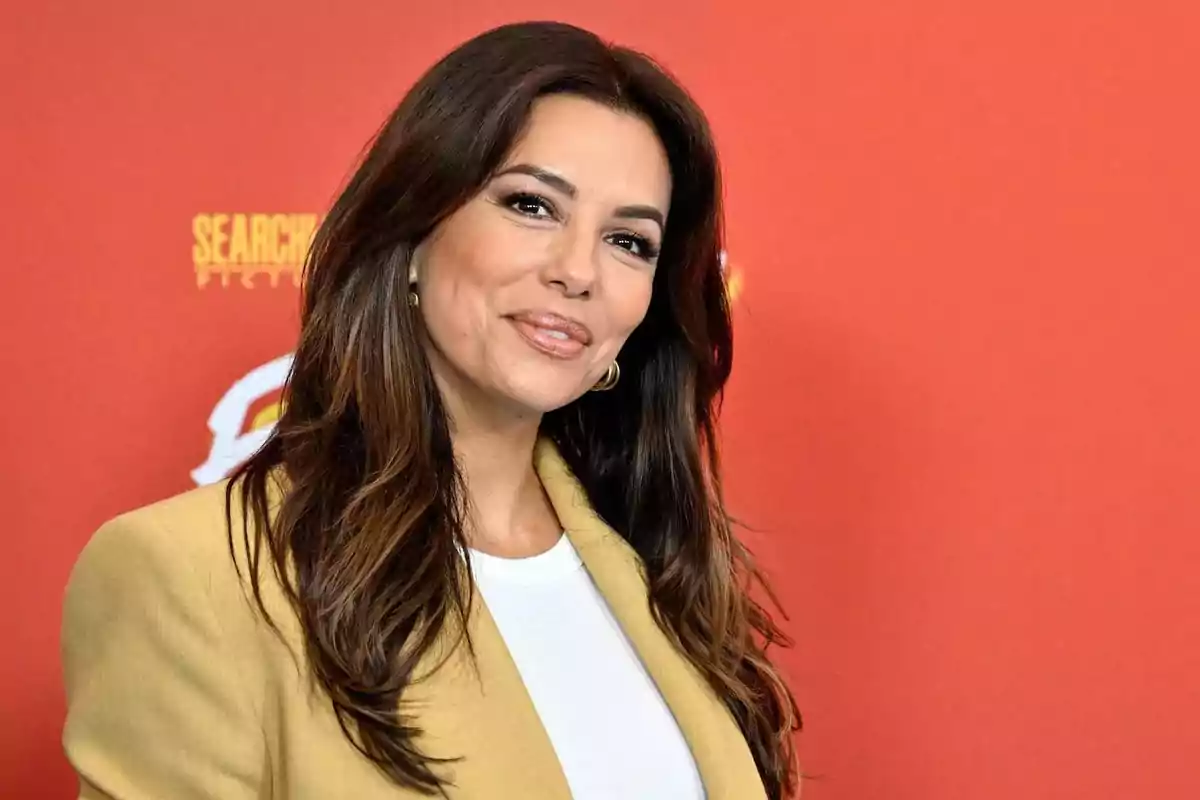 Mujer de cabello largo y oscuro con saco beige y blusa blanca posando frente a un fondo rojo Mujer de cabello largo y oscuro con saco beige y blusa blanca posando frente a un fondo rojo
