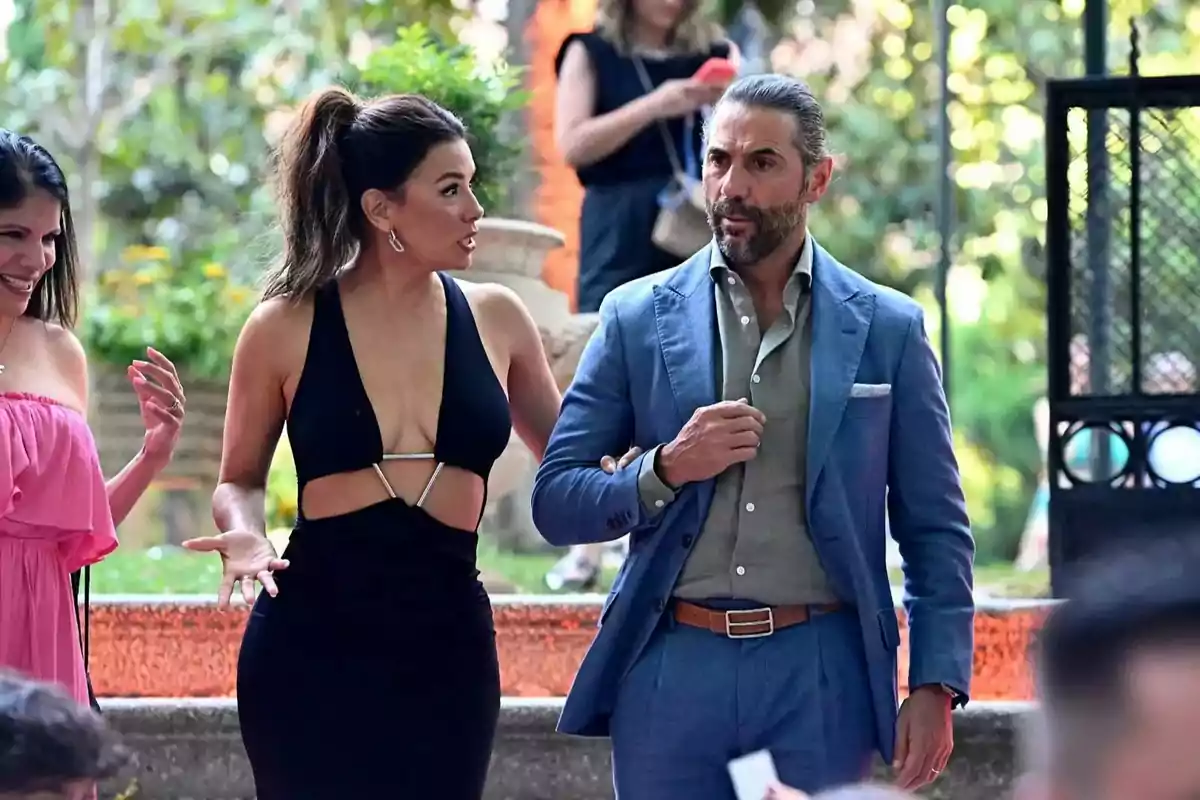 Una mujer con vestido negro elegante y un hombre con traje azul caminan juntos en un evento al aire libre mientras conversan animadamente Una mujer con vestido negro elegante y un hombre con traje azul caminan juntos en un evento al aire libre mientras conversan animadamente